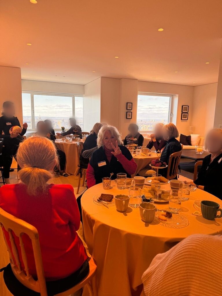 Members gathered around tables set for tea, one smiling and making a peace sign, with large windows overlooking the city behind them